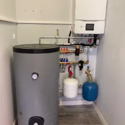 Nowoczesny, biały piec gazowy marki Haier zamontowany nad szarym zasobnikiem ciepłej wody, z widocznymi elementami instalacji hydraulicznymi i grzewczymi w pomieszczeniu z płytkami imitującymi drewno.