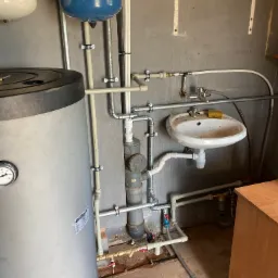 Instalacja wodna z dwoma zbiornikami ciśnieniowymi, bojlerem i umywalką w pomieszczeniu z surowymi ścianami.