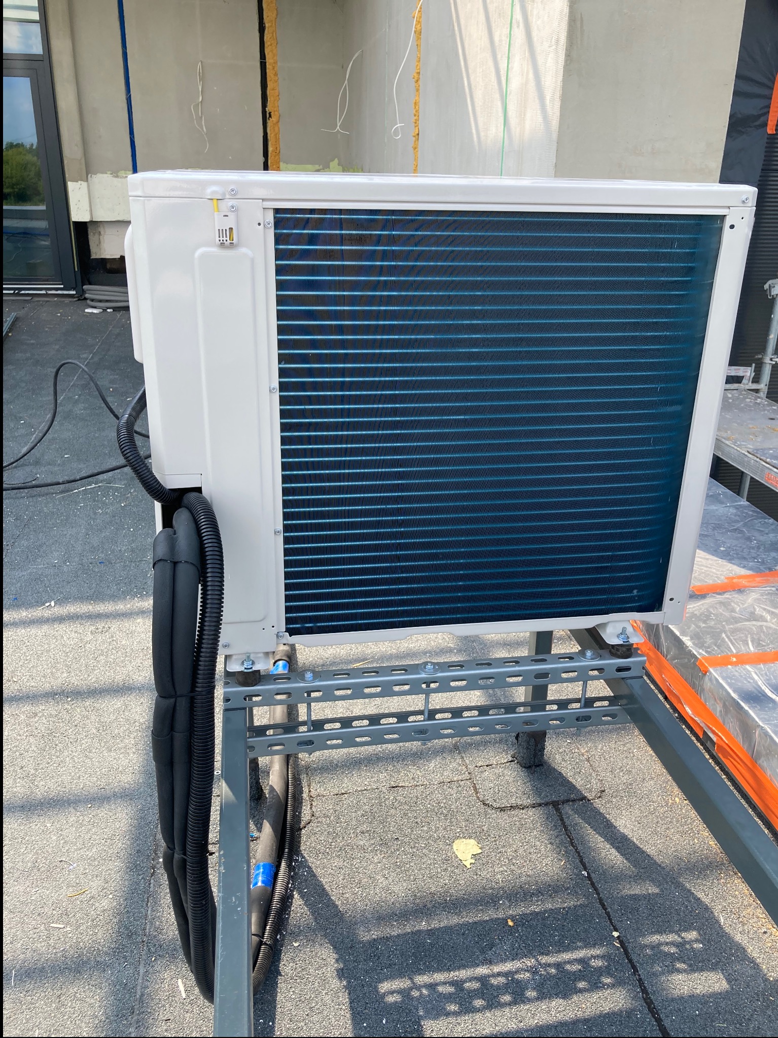 Klimatyzator zewnętrzny na metalowej konstrukcji wsporczej, z izolowanymi rurami podłączonymi z boku, na dachu budynku w trakcie budowy.