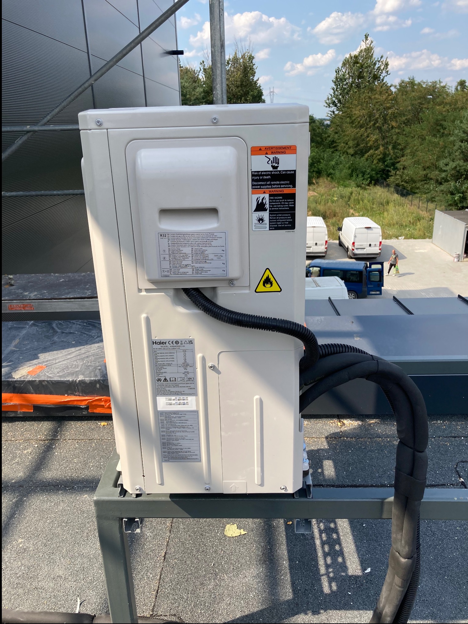Zewnętrzna jednostka klimatyzacji Haier R32 zamontowana na metalowej konstrukcji na dachu, widoczne czarne rury izolacyjne, naklejki ostrzegawcze i informacyjne, w tle budynki i zaparkowane busy.