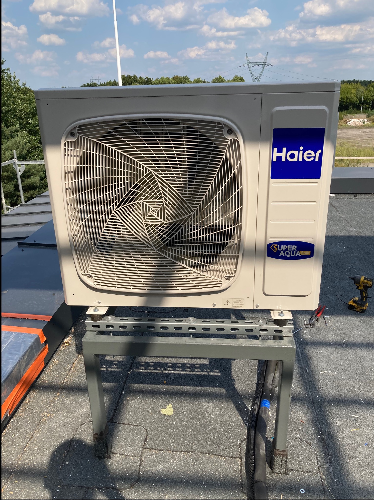 Zewnętrzna jednostka klimatyzacji Haier Super Aqua Split zamontowana na metalowym stelażu na dachu, widoczne narzędzia i przewody, w tle błękitne niebo i słupy wysokiego napięcia.