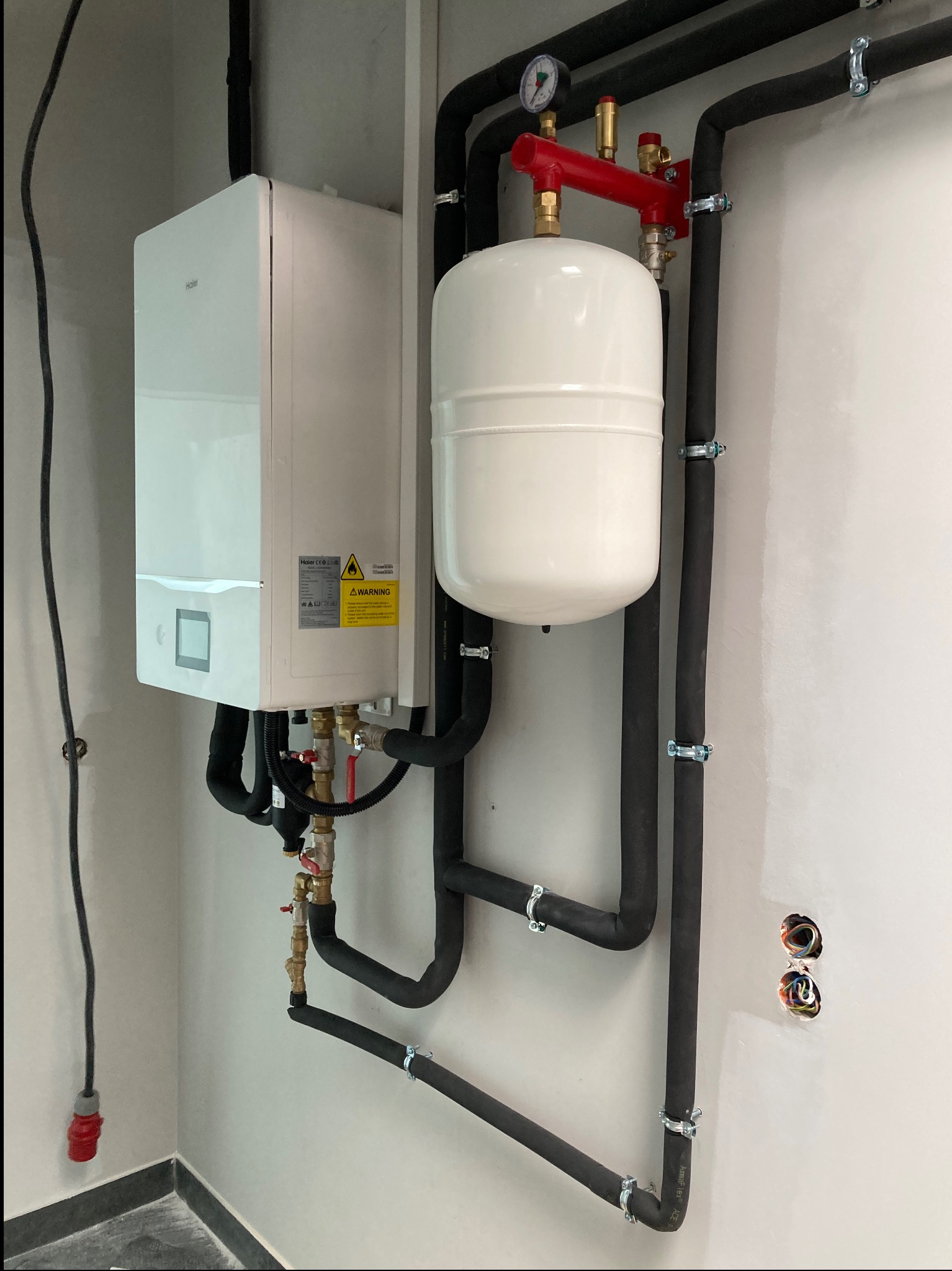 Biały, wiszący piec gazowy marki Haier z podłączonymi czarnymi, izolowanymi rurami, białym zbiornikiem wyrównawczym i czerwonym elementem instalacji, zamontowany na jasnej ścianie z widocznymi...