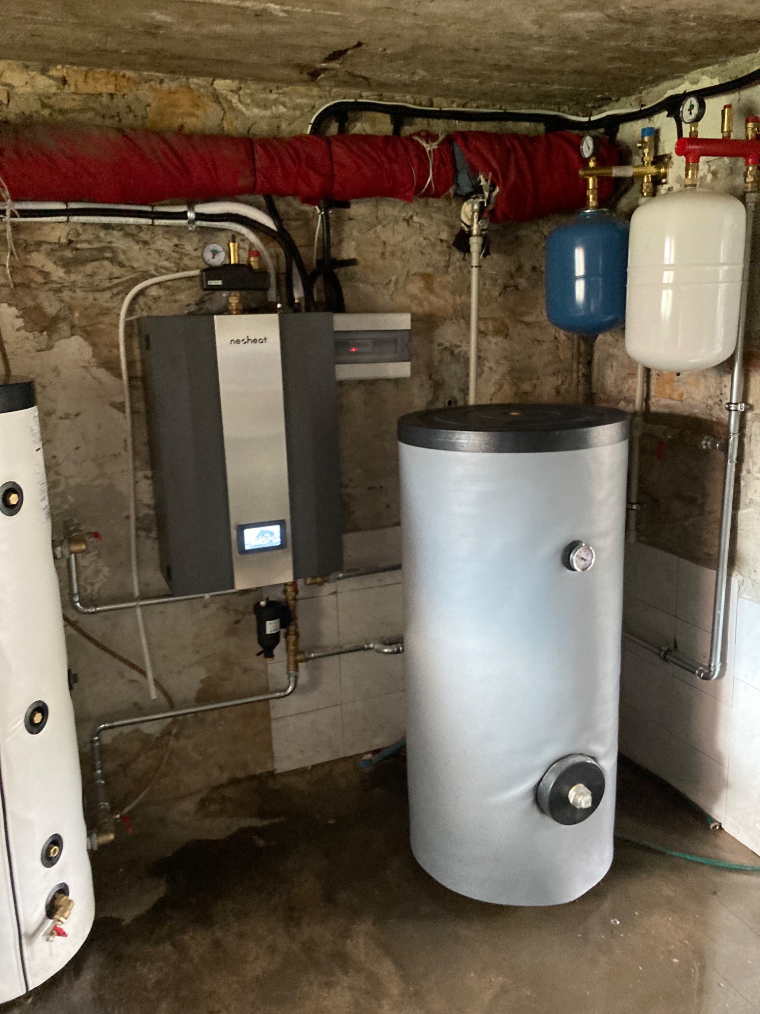 Kotłownia z nowoczesnym szarym piecem Nesheat, dwoma zbiornikami ciśnieniowymi (białym i niebieskim) oraz dużym, srebrnym zasobnikiem ciepłej wody w piwnicy o kamiennych ścianach.