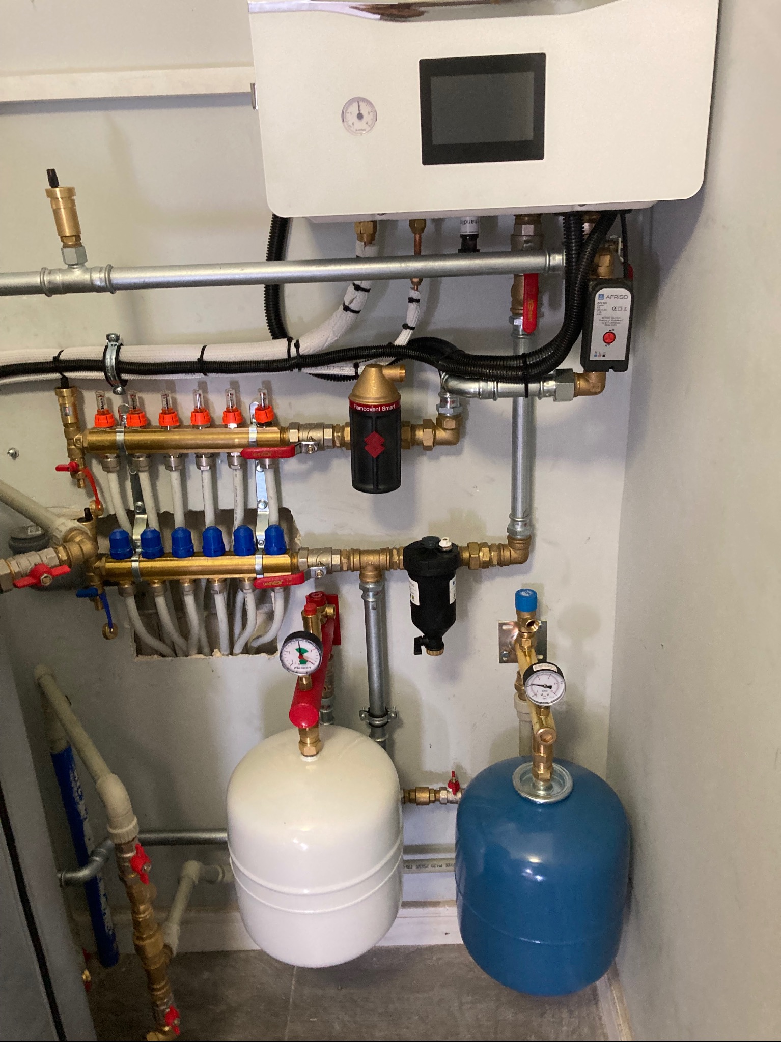 Rozdzielacz instalacji grzewczej z dwoma zbiornikami wyrównawczymi (białym i niebieskim) oraz filtrami, zamontowany na ścianie w pomieszczeniu technicznym.