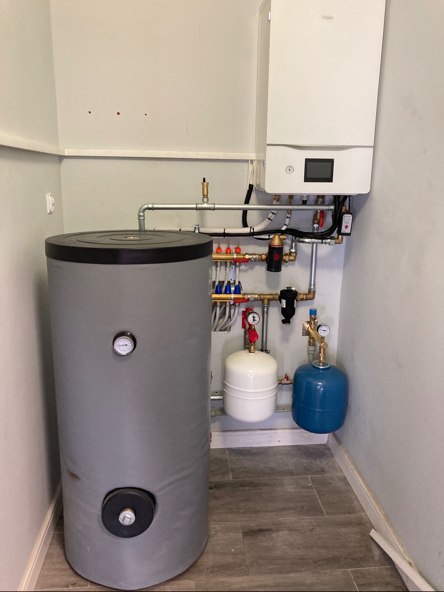 Nowoczesny, biały piec gazowy marki Haier zamontowany nad szarym zasobnikiem ciepłej wody, z widocznymi elementami instalacji hydraulicznymi i grzewczymi w pomieszczeniu z płytkami imitującymi drewno.