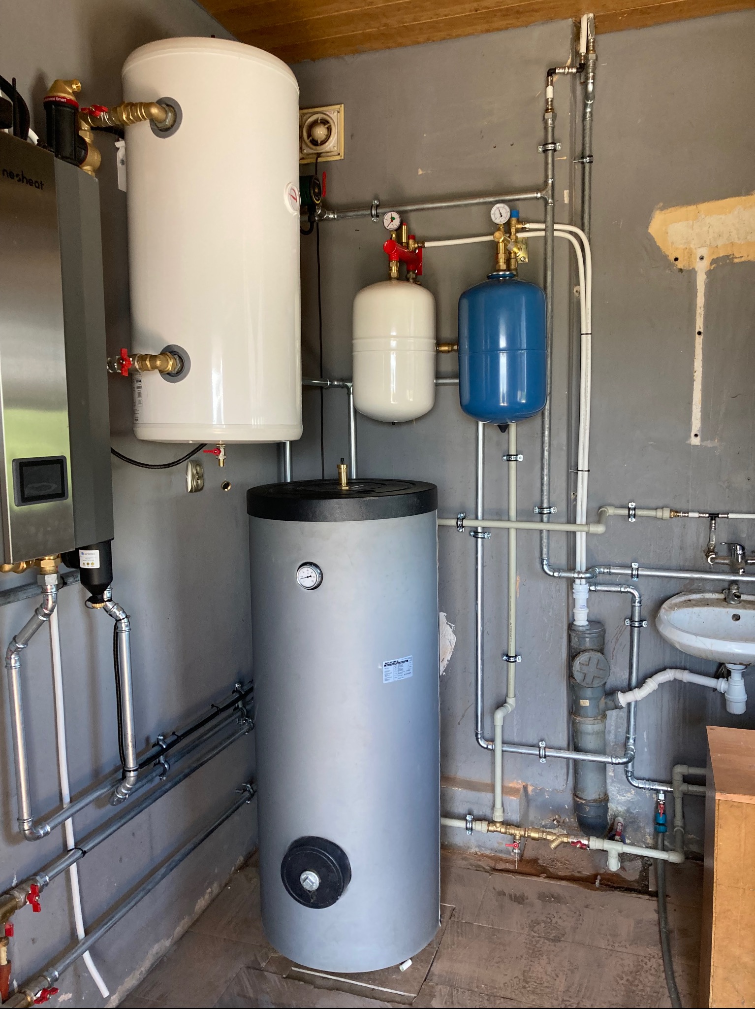 Kotłownia z bojlerami różnej wielkości i koloru, instalacja rurowa z licznymi zaworami i manometrami, szara ściana z widocznymi śladami napraw.