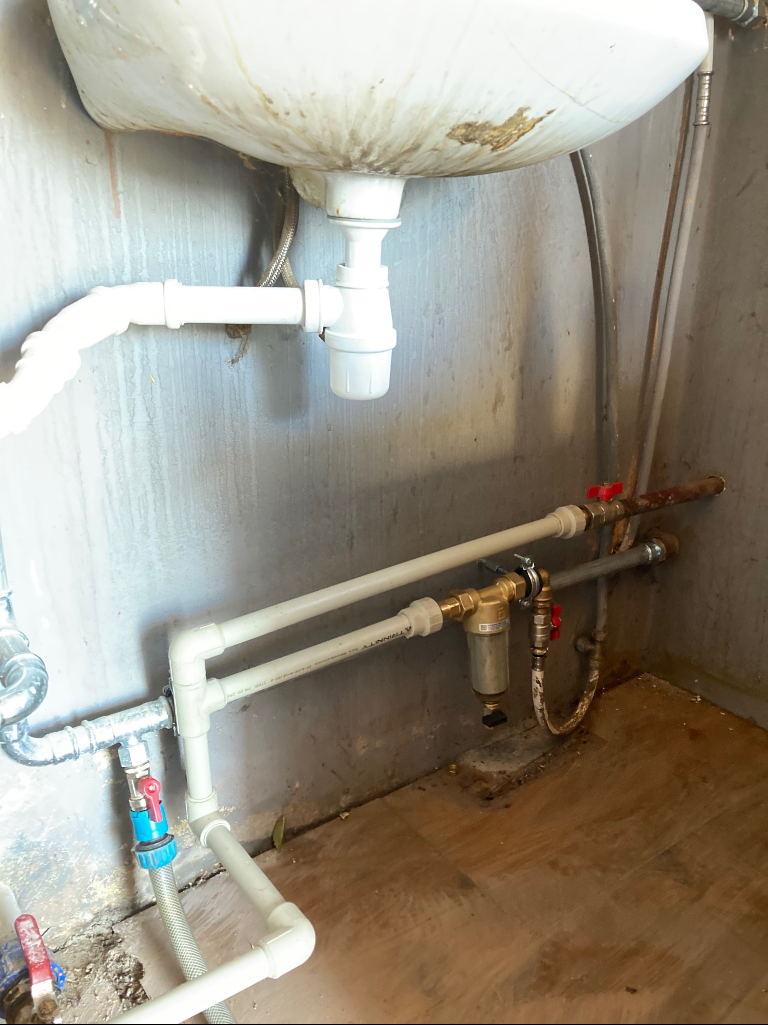 Umywalka z widocznymi instalacjami hydraulicznymi: rury PCV, zawory z czerwonymi uchwytami, filtr wody. Ściany i podłoga wymagają odświeżenia.