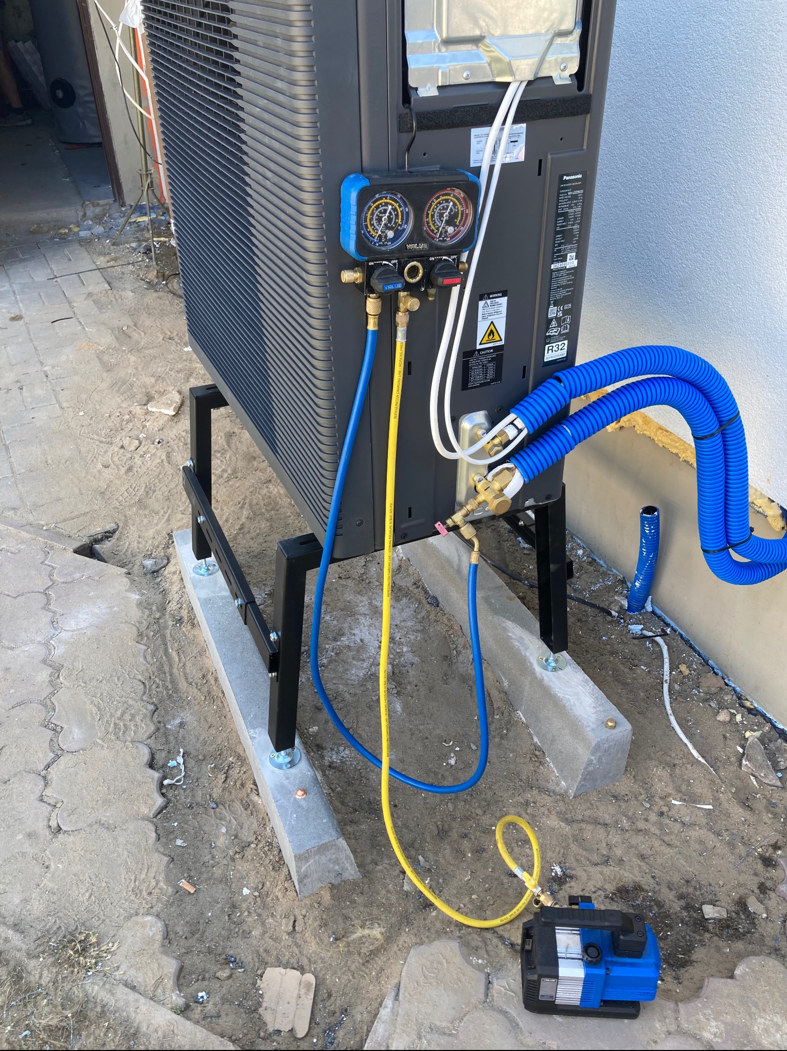Czarna pompa ciepła Panasonic na czarnym metalowym stojaku, podłączona niebieskimi i żółtymi przewodami do manometru i pompy próżniowej, zainstalowana przy białej ścianie budynku.