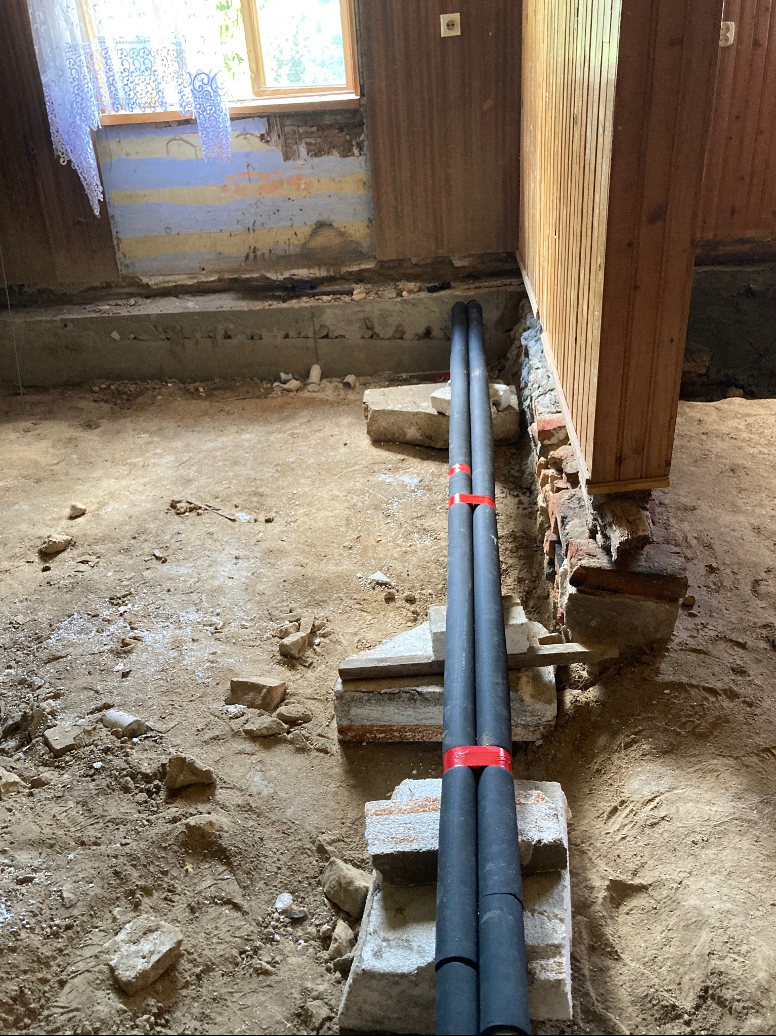 Dwie czarne rury wodne, spięte czerwoną taśmą, prowadzone wzdłuż drewnianej ściany w trakcie prac instalacyjnych w pomieszczeniu z odsłoniętymi fundamentami i resztkami starej tapety.