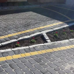 Szara kostka brukowa z żółtymi akcentami i granitowym obramowaniem rabaty z czerwonymi kwiatami. Widok z góry na ułożony wzór.