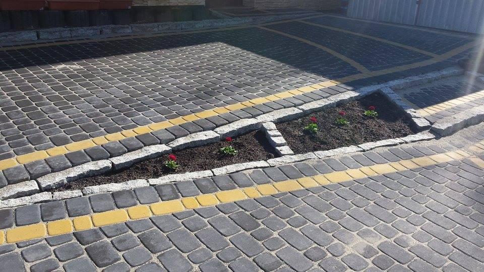 Szara kostka brukowa z żółtymi akcentami i granitowym obramowaniem rabaty z czerwonymi kwiatami. Widok z góry na ułożony wzór.