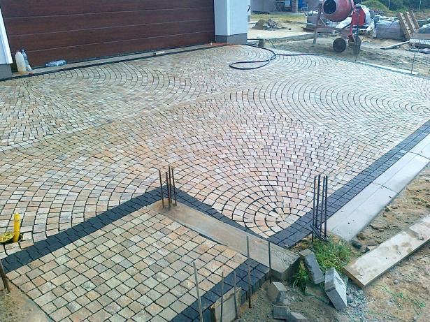Ułożona kostka granitowa w kształcie wachlarza przed garażem. Widoczne obrzeża z ciemnej kostki i elementy przygotowane do dalszych prac.