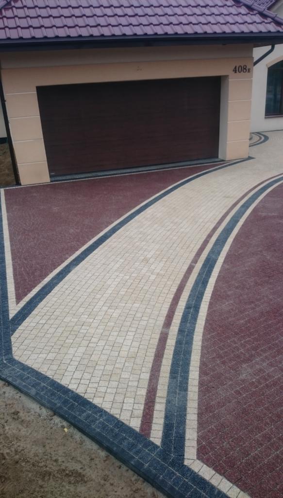 Podjazd z kostki brukowej w odcieniach czerwieni i bieli, z dekoracyjnymi pasami w kolorze granatowym, prowadzący do garażu domu z beżową elewacją.