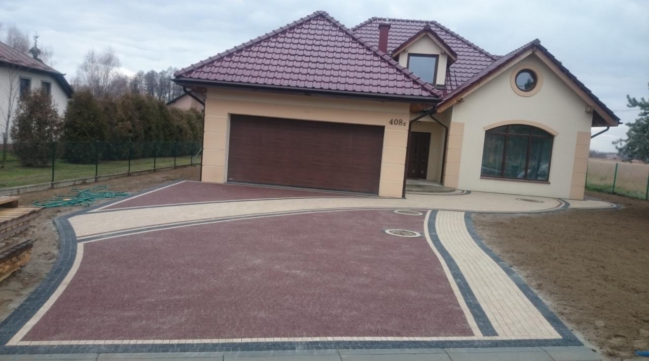 Nowoczesny podjazd z kostki brukowej w odcieniach brązu i beżu, prowadzący do domu z garażem. Widoczne studzienki kanalizacyjne i elementy dekoracyjne z kostki.