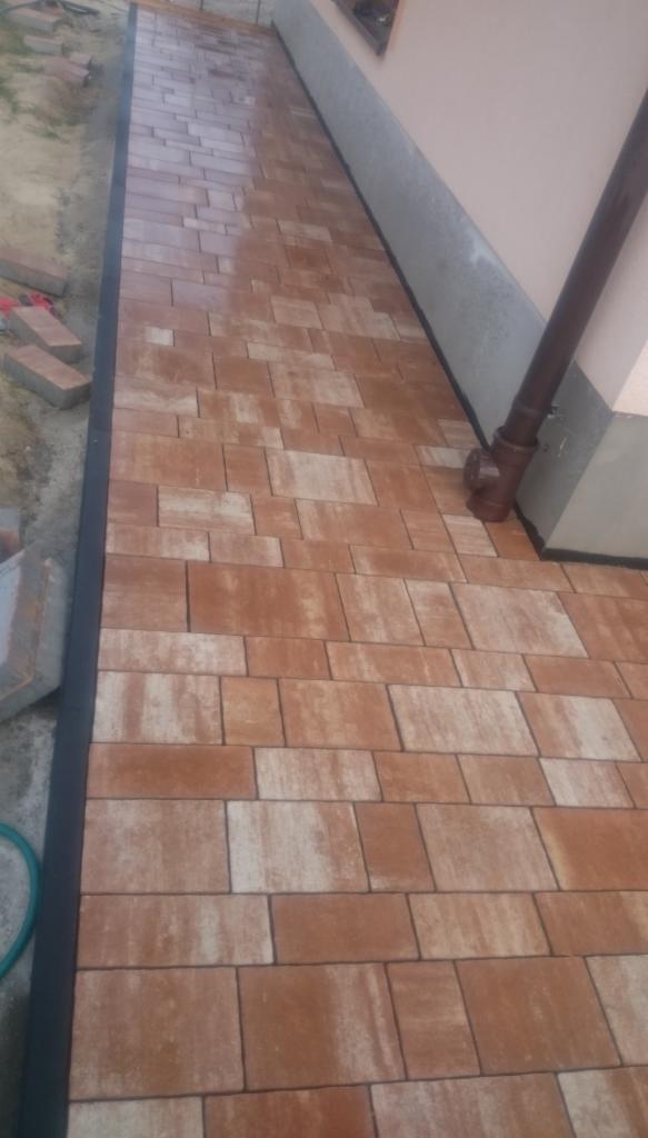 Ułożona kostka brukowa w odcieniach brązu i beżu, przylegająca do ściany budynku z rynną. Częściowo widoczne kostki do ułożenia.