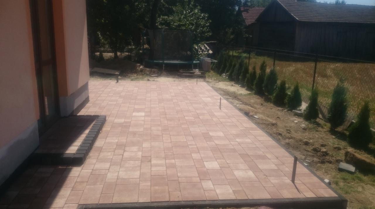 Nowa, beżowa kostka brukowa ułożona na posesji, widok z boku domu. Trampolina w tle. Ciemne obrzeże kostki.
