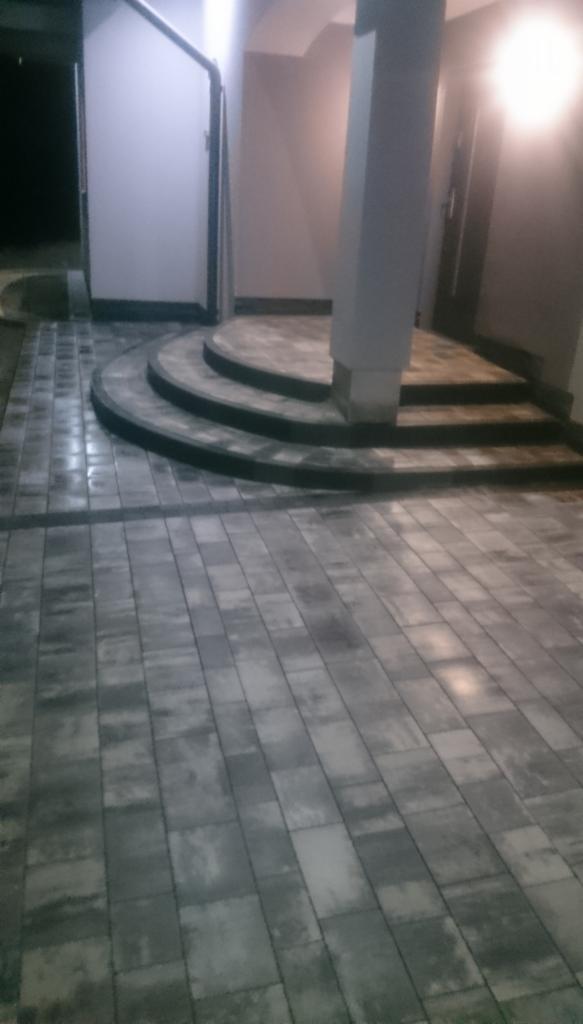 Szara kostka brukowa ułożona na podjeździe z trzema stopniami. Na górze kolumna i drzwi wejściowe. Widok z dołu na zewnątrz budynku.