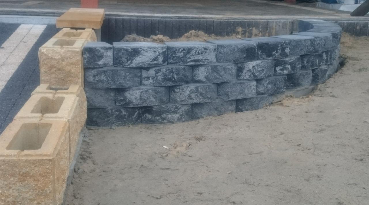 Mur oporowy z szarych, betonowych bloków ułożonych łukiem, połączony z prostopadłą ścianą z beżowych pustaków. Widoczny piasek i fragment chodnika.