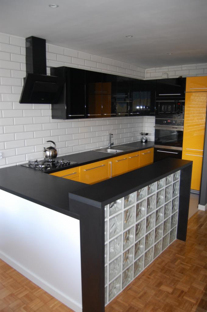 Nowoczesna kuchnia z czarnymi szafkami, żółtymi akcentami i wyspą z luksferami. Minimalistyczny design, białe płytki na ścianie, drewniana podłoga.