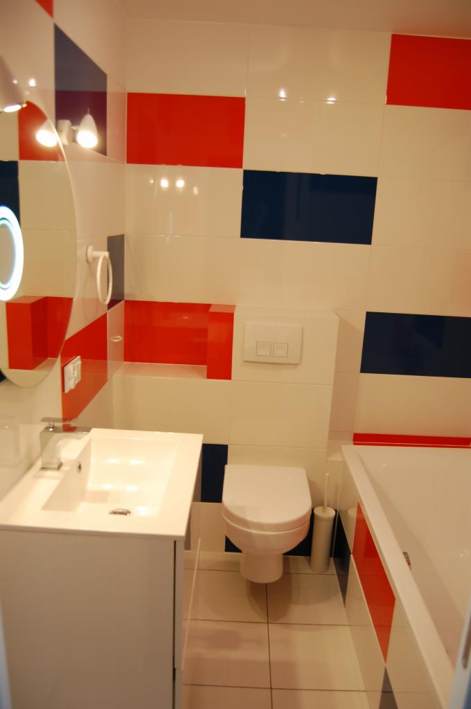 Wnętrze łazienki z białą umywalką, toaletą i wanną. Ściany wyłożone płytkami w kolorach białym, czerwonym i granatowym, tworząc geometryczny wzór.
