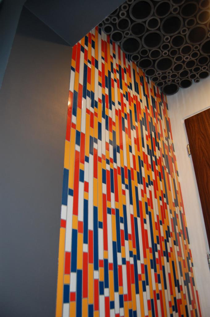 Wnętrze z dekoracyjną ścianą z kolorowych, pionowych pasków (czerwony, żółty, biały, niebieski) i sufitem z okrągłych elementów. Widoczny fragment drzwi.