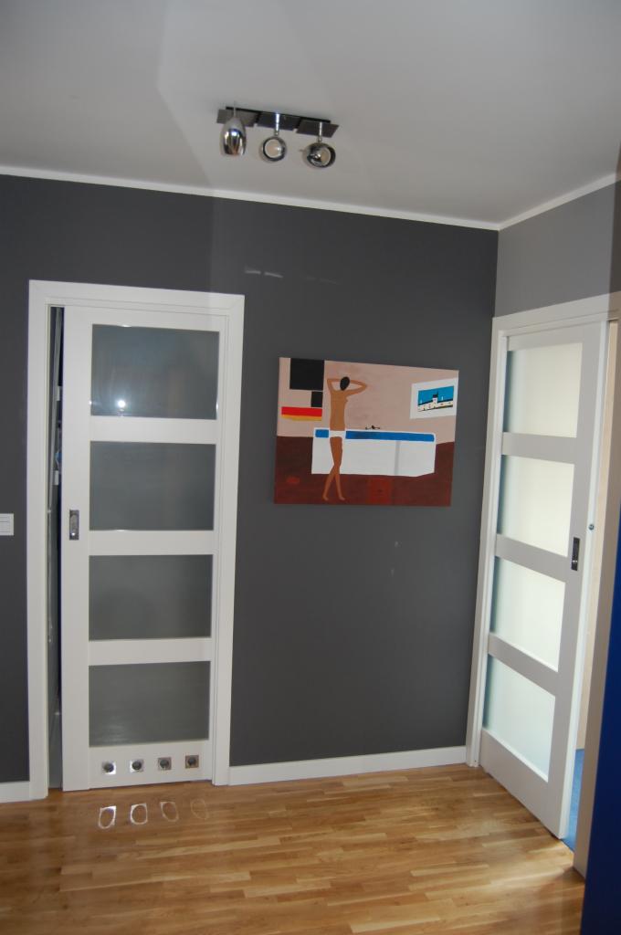 Nowoczesne, białe drzwi przesuwne z czterema panelami szklanymi, zamontowane w szarej ścianie nad parkietem. Nad drzwiami widoczny obraz i lampa sufitowa.