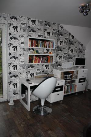 Pokój młodzieżowy z biurkiem i regałem, ściana w komiksowe wzory. Białe meble, drewniana podłoga. Nietypowy układ pomieszczenia na poddaszu.