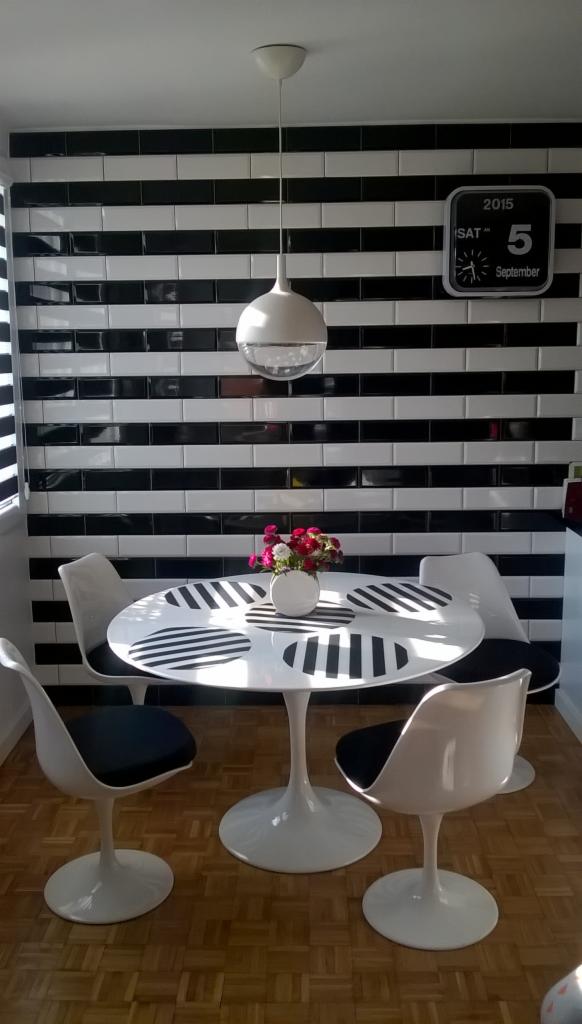 Wnętrze jadalni w czarno-białej tonacji: okrągły stół z bukietem, krzesła tulipanowe, ściana w pasy z płytek i zegar z datą 5 września 2015.