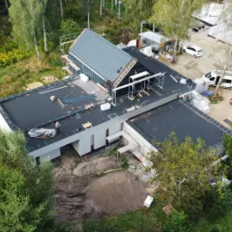 Widok z lotu ptaka na budowę domu jednorodzinnego z płaskim dachem pokrytym czarną papą i częściowo ukończonym dachem dwuspadowym z szarej blachy, otoczonego lasem i z zaparkowanymi samochodami w tle.