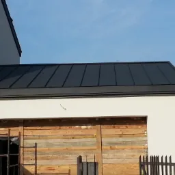 Nowoczesny, grafitowy dach z blachy na budynku w trakcie budowy, z deskami zasłaniającymi otwór garażowy.