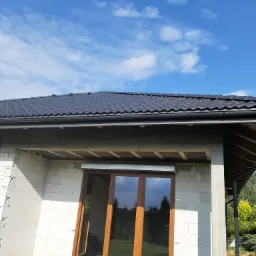 Czarny dach z dachówki ceramicznej, nowo wybudowany dom z pustaków, okna z brązowymi ramami i błękitne niebo w tle.