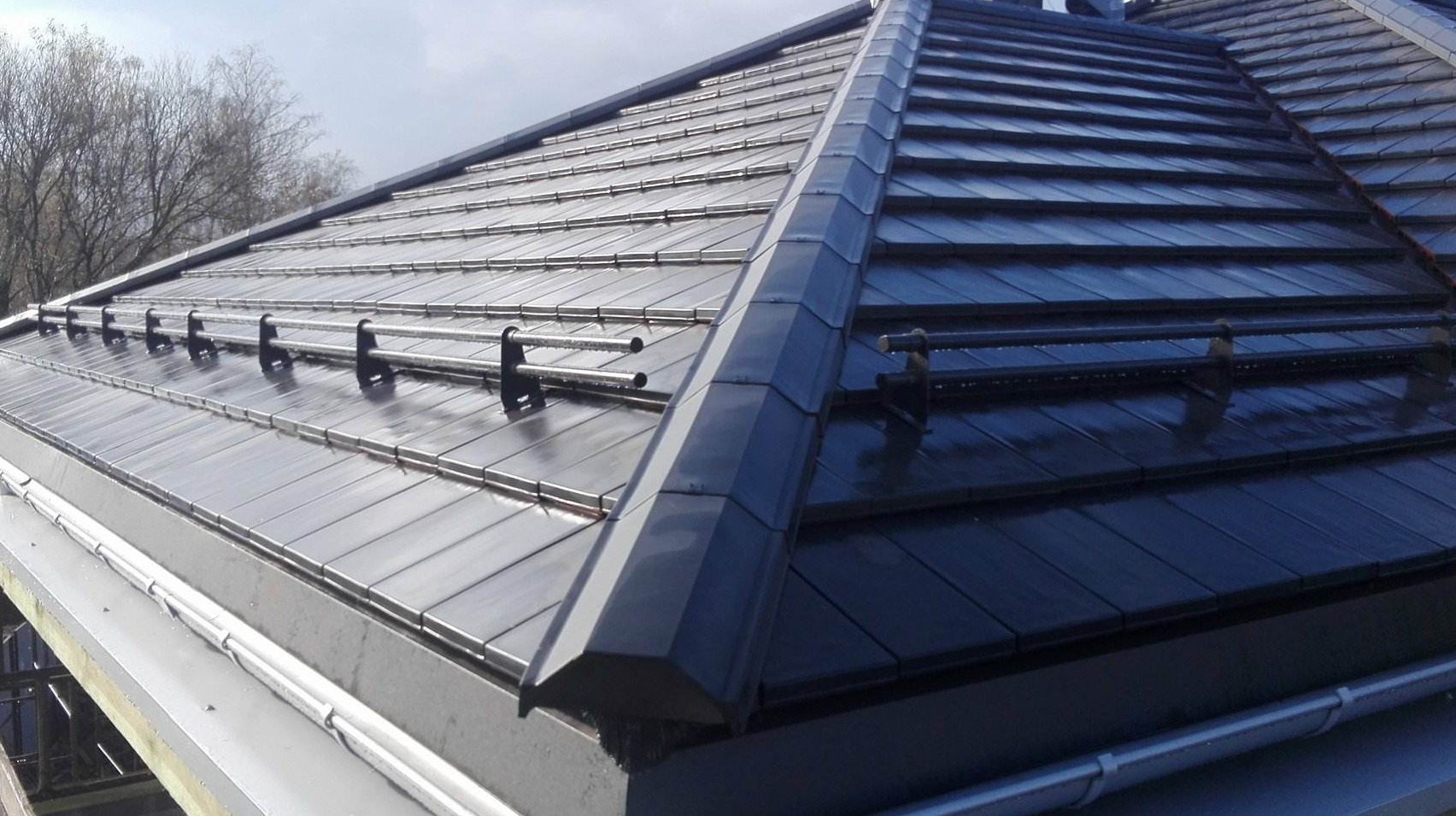 Ciemna, błyszcząca dachówka ceramiczna na nowym dachu z zamontowanymi rurami odgromowymi i orynnowaniem, widok z dołu.