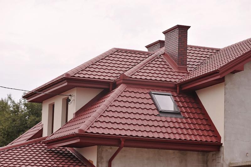 Czerwony dach z dachówki metalowej z oknem dachowym i ceglanym kominem, widoczne rynny w tym samym kolorze, fragment budynku w stanie surowym.