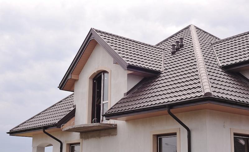 Dom w trakcie budowy z nowym, brązowym dachem z blachy profilowanej, widoczne rynny i komin wentylacyjny.