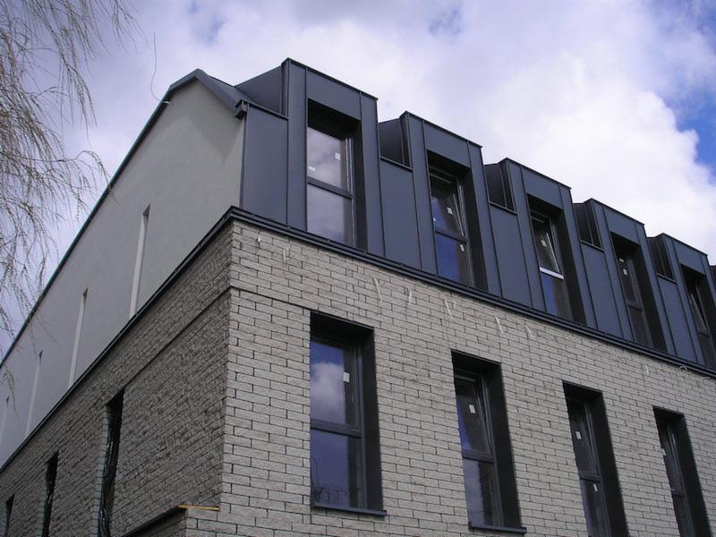 Nowoczesny budynek z szarej cegły z ciemnymi oknami i dachem z lukarnami pokrytym metalem w kolorze grafitowym, widok z dołu pod kątem.