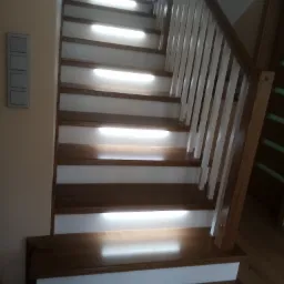 Drewniane schody z białymi podstopnicami i oświetleniem LED pod każdym stopniem. Balustrada drewniana z białymi tralkami. Widok z dołu, lekko z boku.