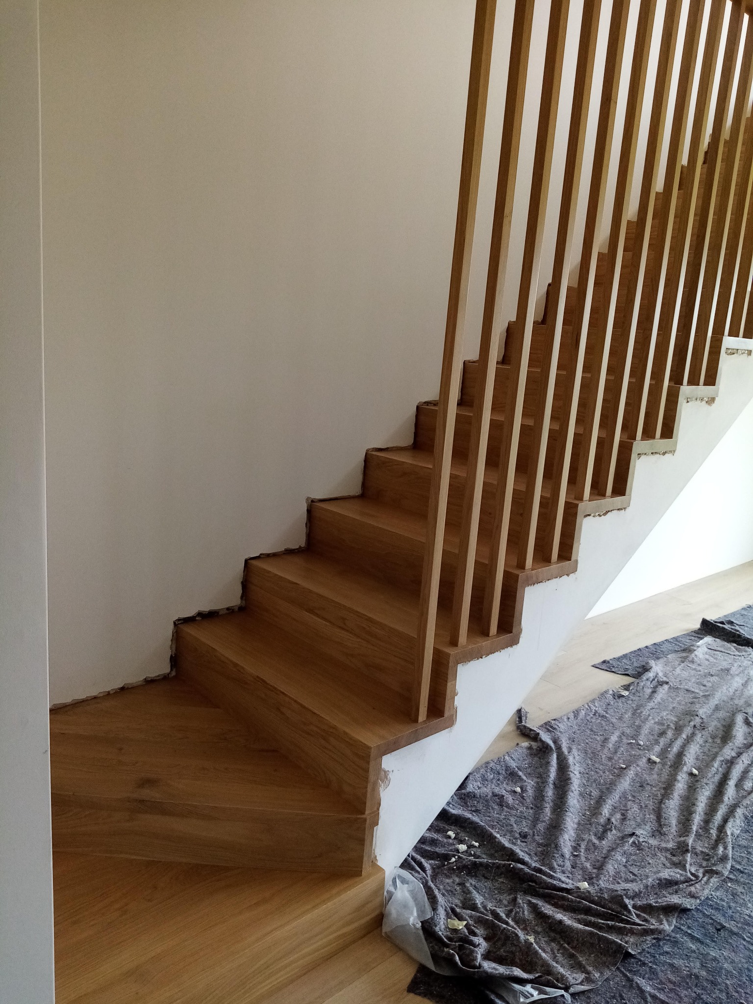 Jasne, drewniane schody z nowoczesną balustradą z pionowych listew. Widoczne zabezpieczenia podłogi. Ujęcie z dołu, ukazujące detale wykończenia.