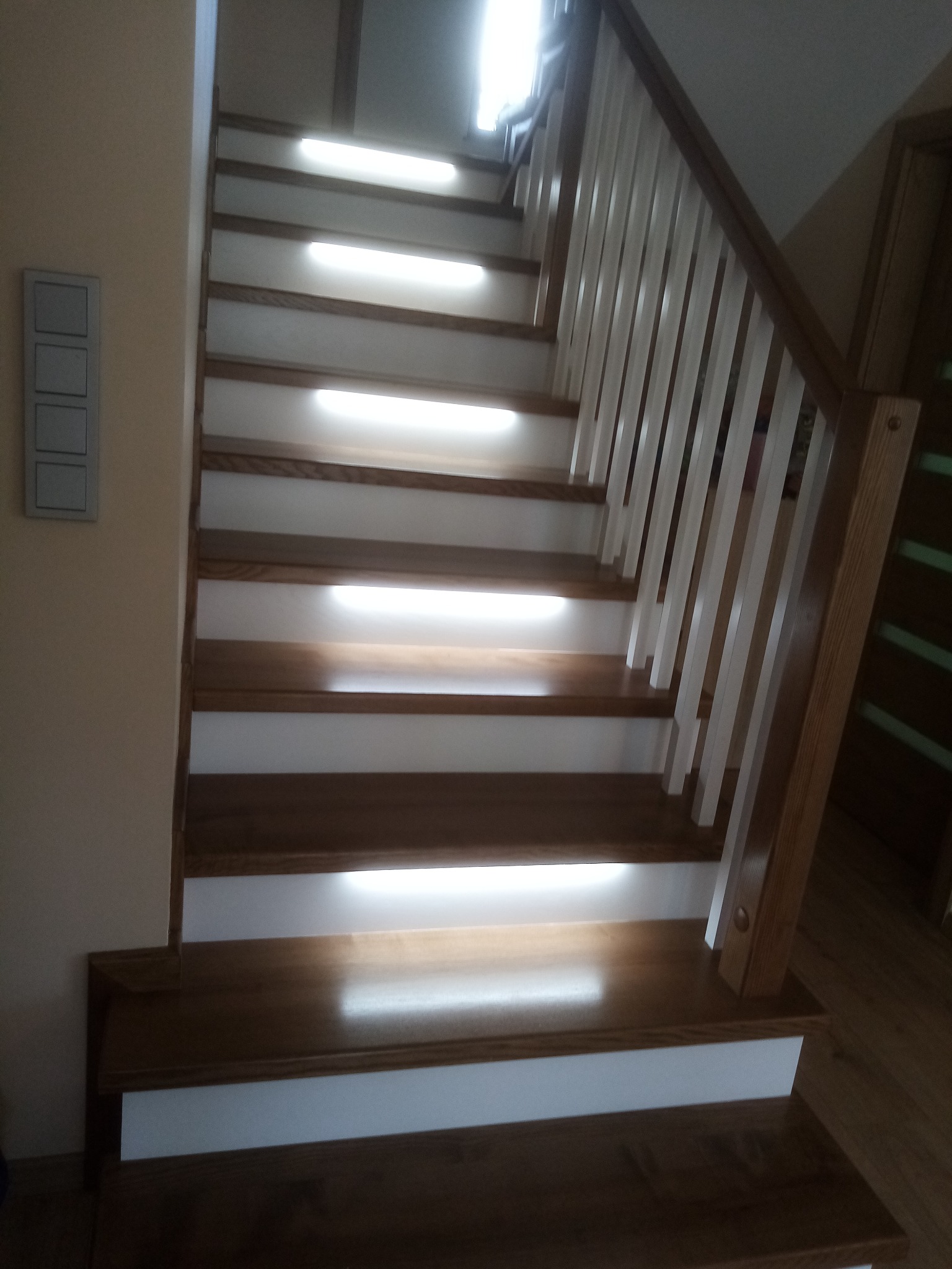 Drewniane schody z białymi podstopnicami i oświetleniem LED pod każdym stopniem. Balustrada drewniana z białymi tralkami. Widok z dołu, lekko z boku.