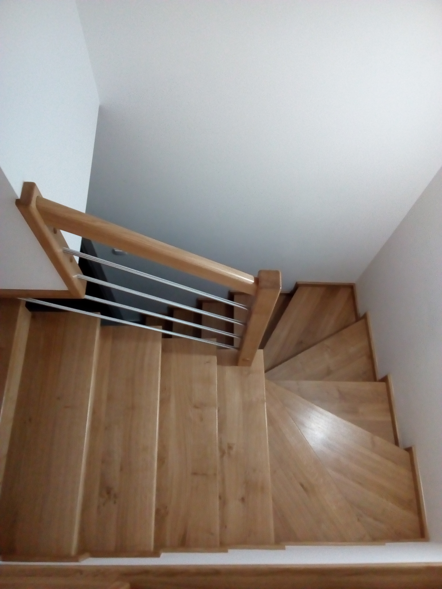 Widok z góry na drewniane schody z jasnego drewna, z balustradą z drewnianą poręczą i metalowymi prętami, prowadzące w dół wzdłuż białej ściany.