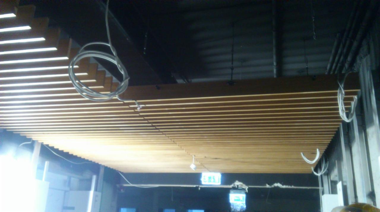 Drewniany sufit lamelowy z widocznymi przewodami elektrycznymi i oświetleniem awaryjnym, w trakcie montażu.