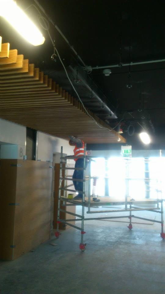 Pracownik w kamizelce odblaskowej na rusztowaniu montuje drewniane lamele sufitowe w pomieszczeniu z czarnym sufitem i oświetleniem punktowym.