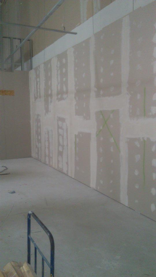 Ściana z płyt kartonowo-gipsowych w trakcie montażu, widoczne łączenia wzmocnione taśmą, metalowe profile sufitowe i zielone oznaczenia na ścianie.