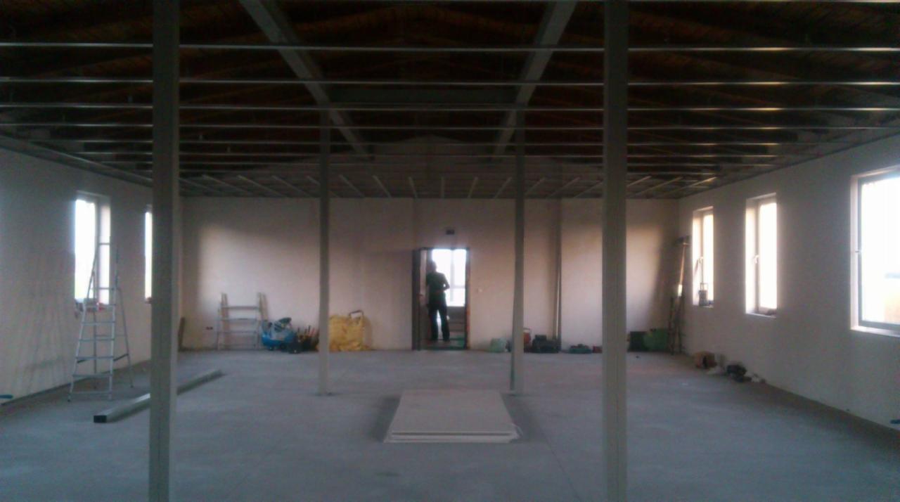 Wnętrze w trakcie prac wykończeniowych, widoczne metalowe profile sufitowe, ściany przygotowane pod malowanie, osoba w przejściu drzwiowym, drabiny i narzędzia w tle.