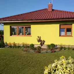 Żółty dom jednorodzinny z czerwonym dachem i zadbanym trawnikiem oraz rabatami kwiatowymi, widok z przodu w słoneczny dzień.