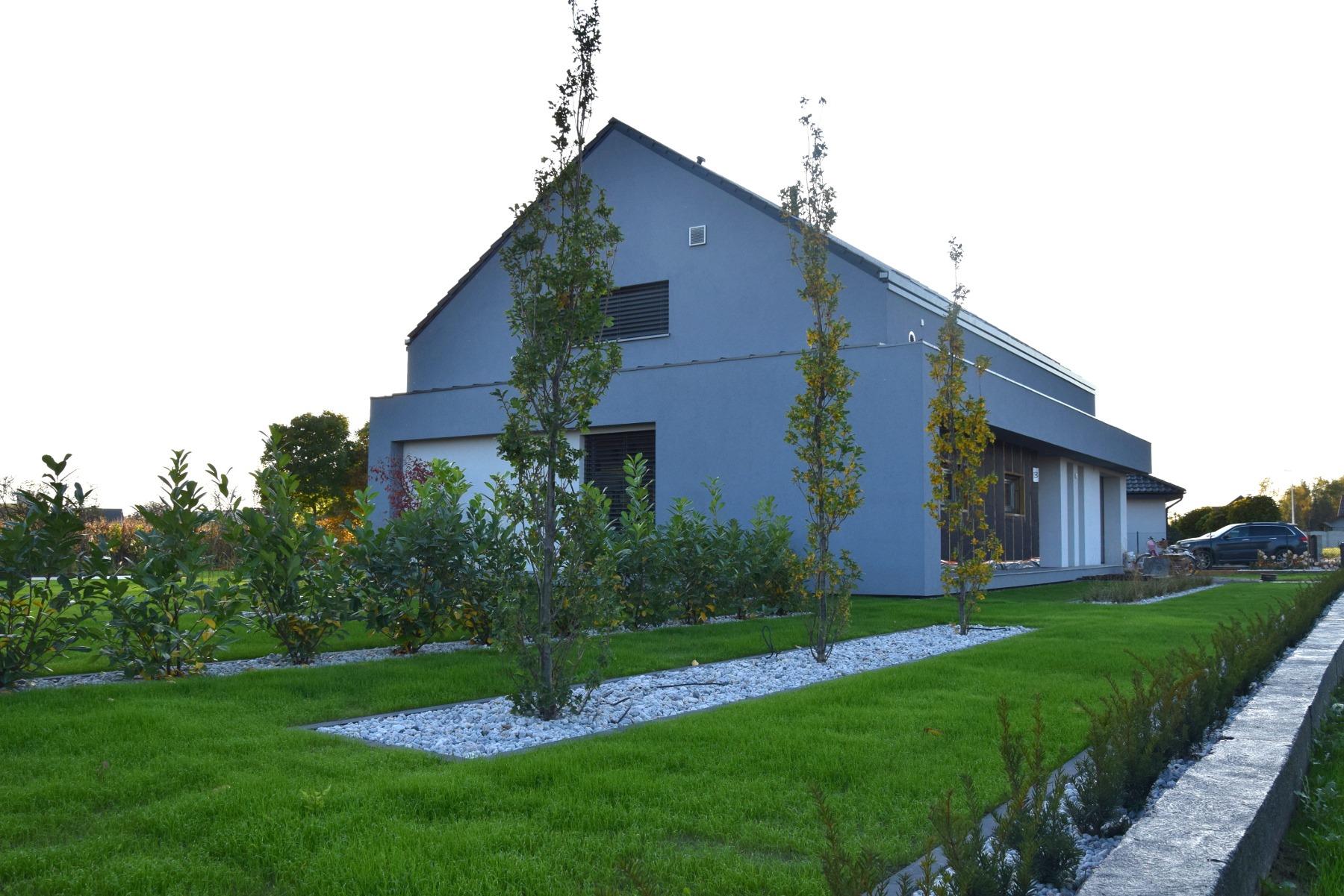 Nowoczesny dom z szarą elewacją, otoczony świeżo założonym trawnikiem, rabatami z białym grysem i młodymi drzewkami.