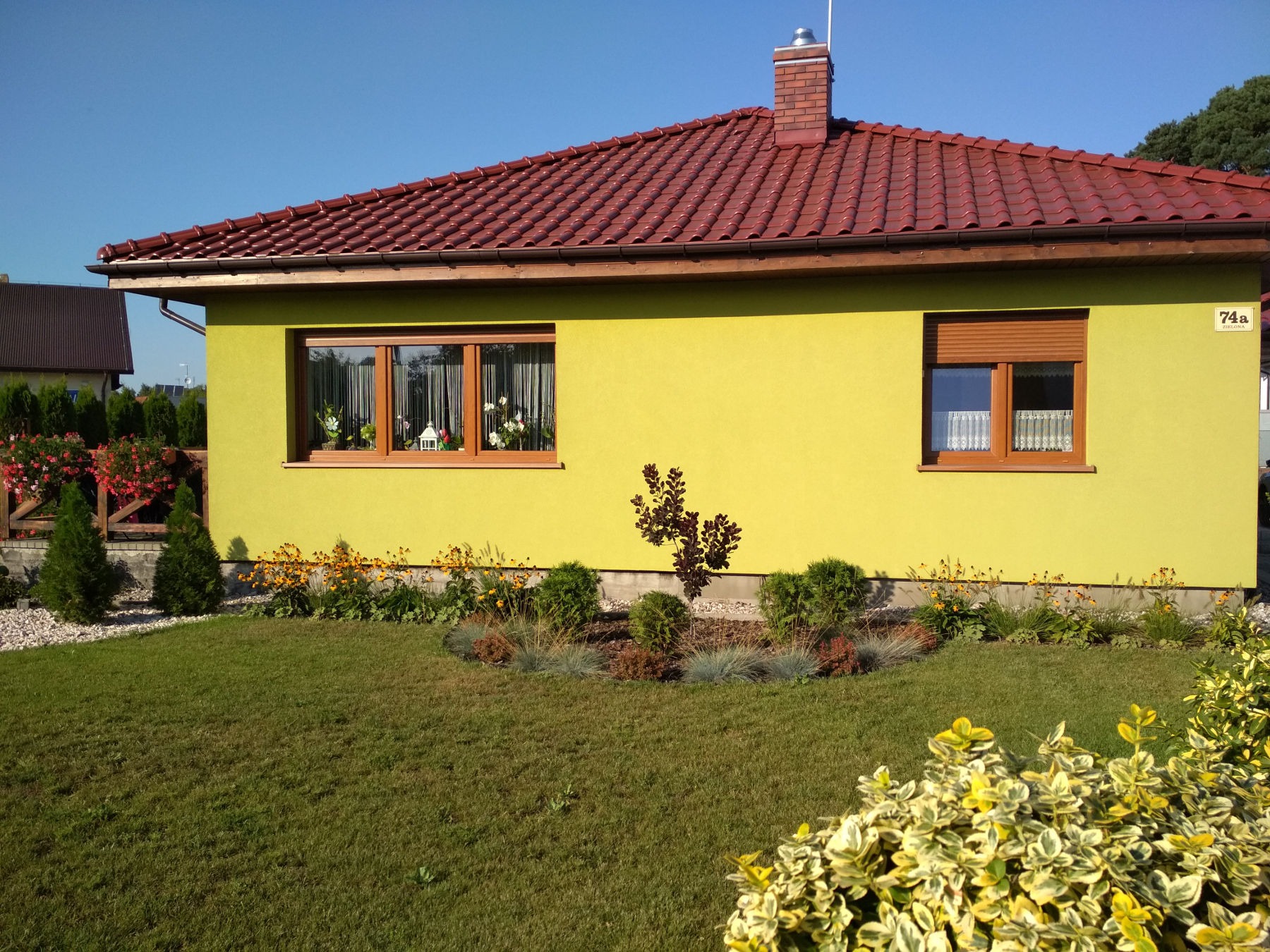 Żółty dom jednorodzinny z czerwonym dachem i zadbanym trawnikiem oraz rabatami kwiatowymi, widok z przodu w słoneczny dzień.