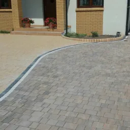 Ułożona kostka brukowa w dwóch odcieniach szarości i beżu tworząca ścieżkę i podjazd przed domem z ceglaną elewacją i kwiatami w donicach na schodach.