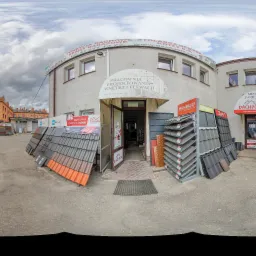 Ekspozycja różnych rodzajów pokryć dachowych przed budynkiem firmy z branży budowlanej, widoczne dachówki ceramiczne i blaszane w różnych kolorach i wzorach, z przodu budynku reklamy firm i produktów.