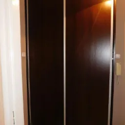 Ciemna szafa wnękowa z przesuwnymi drzwiami i srebrnymi obramowaniami, widoczna w korytarzu, oświetlona lampą sufitową. Minimalistyczny design.