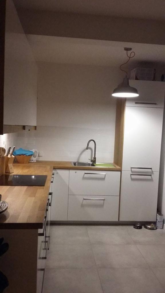 Nowoczesna kuchnia z drewnianym blatem i białymi frontami szafek. Nad zlewem wisi lampa w stylu industrialnym. Widoczne płytki na ścianie i akcesoria kuchenne.